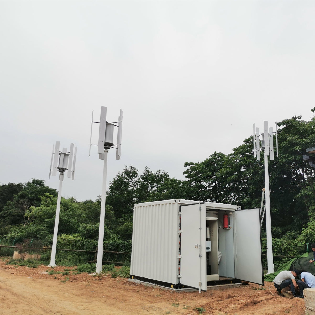 H series vertical wind turbines with the centre assist wheel at a remote site beside an equipment container — the turbine without a centre drum is a different model