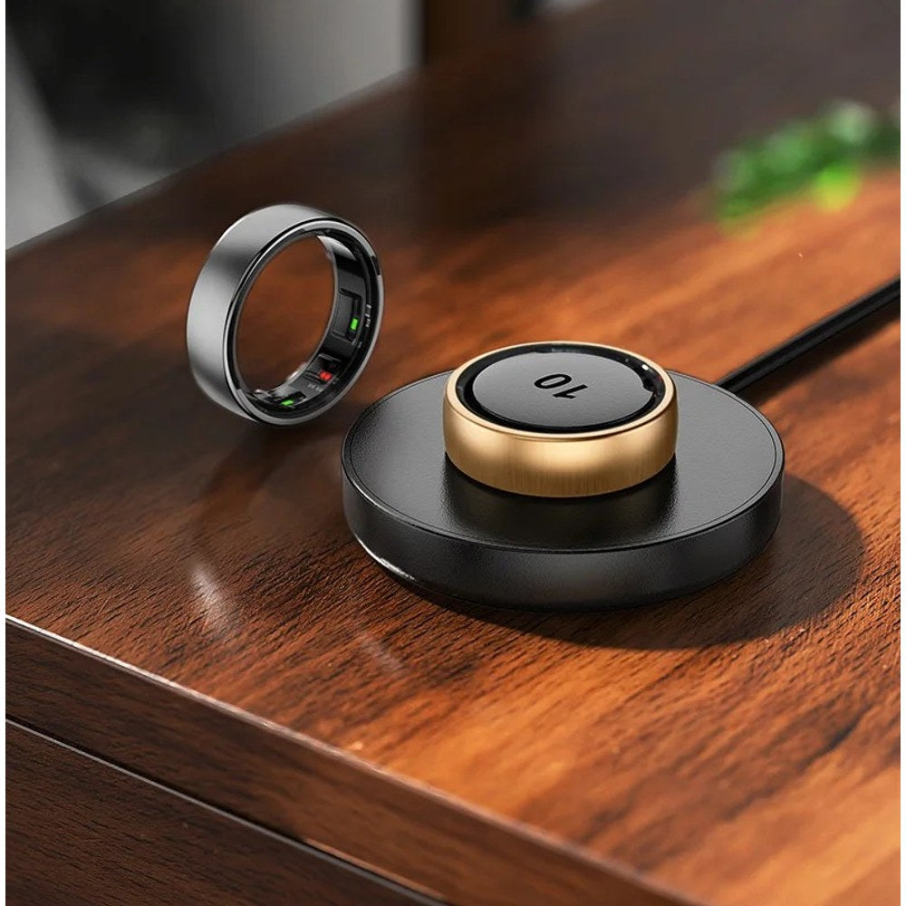 Gold R10 ring seated on the round black charging dock with its lead attached, silver ring beside it on a timber table