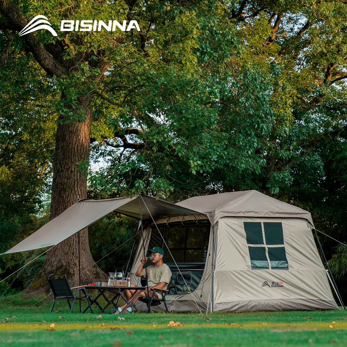 Sand BISINNA cabin tent pitched under a tree with the canopy extended, camper seated at a table.