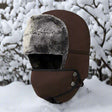 Brown trapper hat side on, grey pile lining showing, face panel fitted, snowy branches