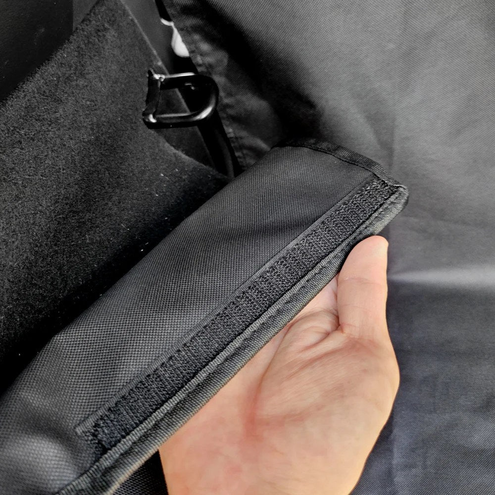 Hand holding the edge of a liner flap showing the hook-and-loop strip sewn along it, inside a car cargo area
