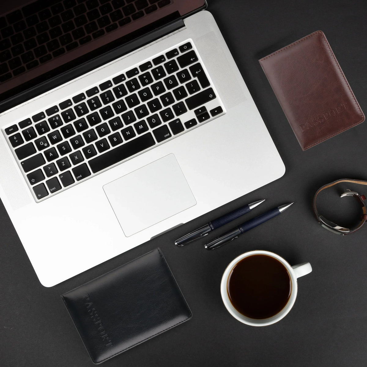 Coffee and black passport covers laid on a desk beside a laptop, pens and a coffee cup