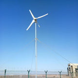 Three-blade turbine on a guyed steel mono-pole against clear blue sky, inside a fenced compound with a concrete building behind.