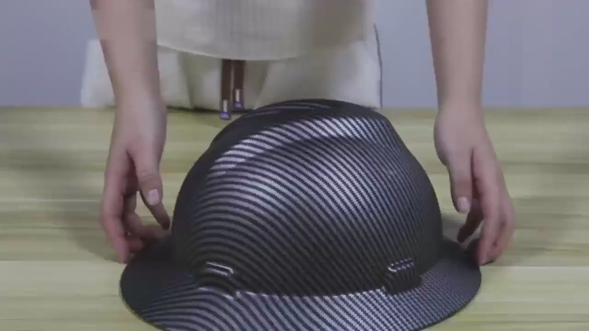 Black carbon-pattern full-brim hard hat on a bench: hands turn it over to show the harness, the headband and the adjuster inside.
