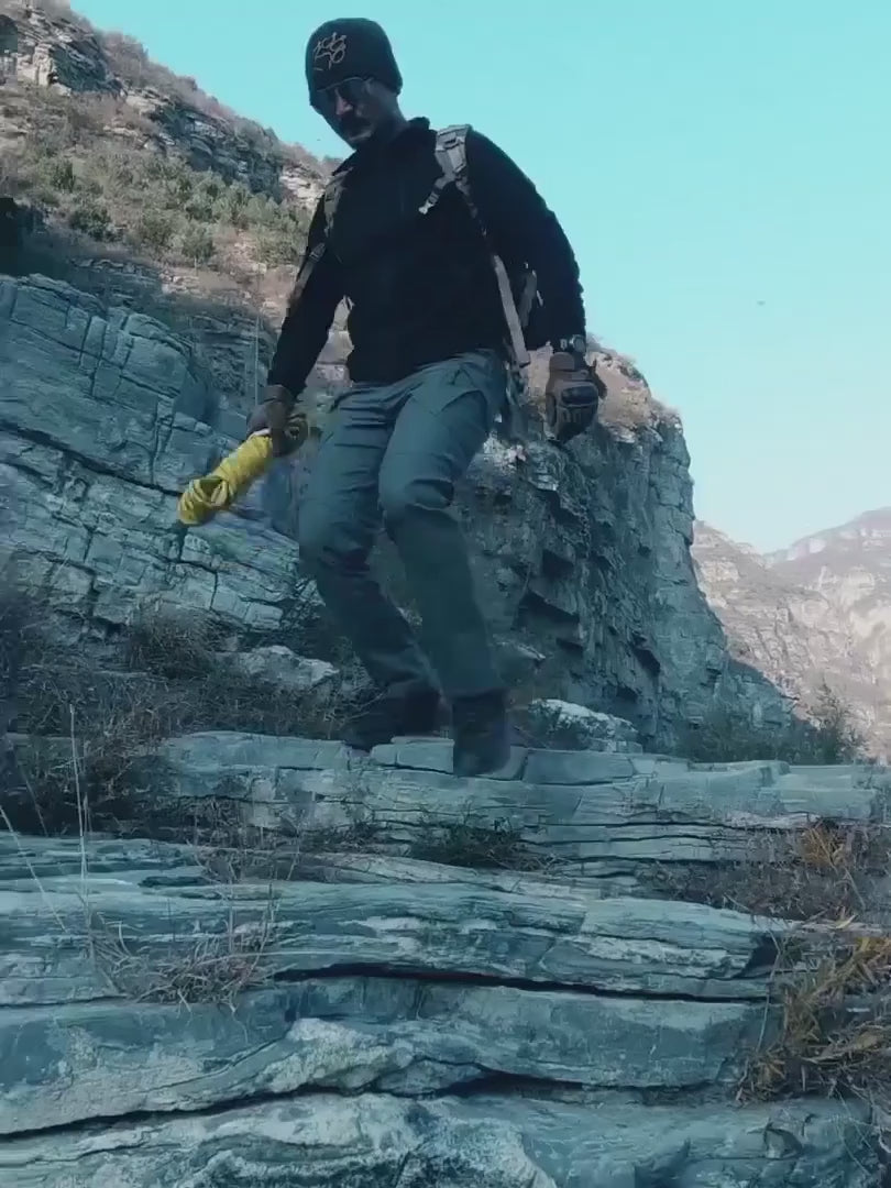 Man in grey cargo trousers climbing rocks and crossing a creek, with a close-up of a zipped thigh pocket and the knees bending.