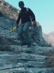 Man in grey cargo trousers climbing rocks and crossing a creek, with a close-up of a zipped thigh pocket and the knees bending.