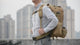 Man on a rooftop wearing the khaki 4-in-1 MOLLE pack with its front waist pack and two side pouches attached, turning to show the back, the front harness and the waist strap.
