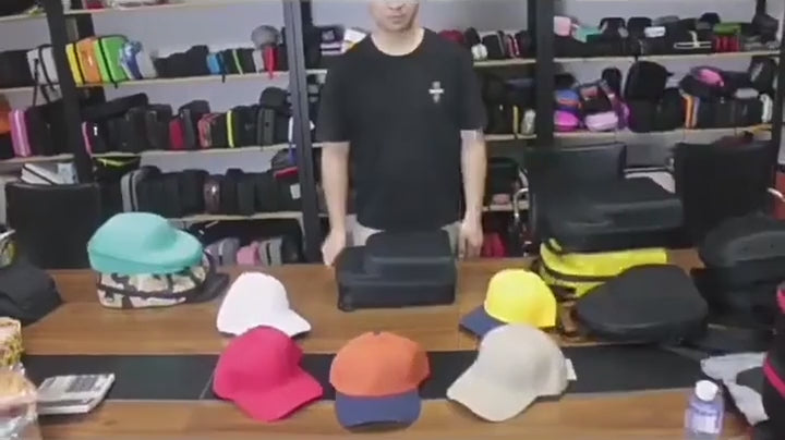 A man packing several baseball caps into the black moulded cap case on a workbench, zipping it shut and carrying it.