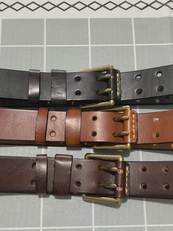 Three 38 mm leather belts with double prong buckles on a grid mat; gloved hands thread one through its keeper and run along the strap.
