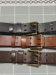 Three 38 mm leather belts with double prong buckles on a grid mat; gloved hands thread one through its keeper and run along the strap.