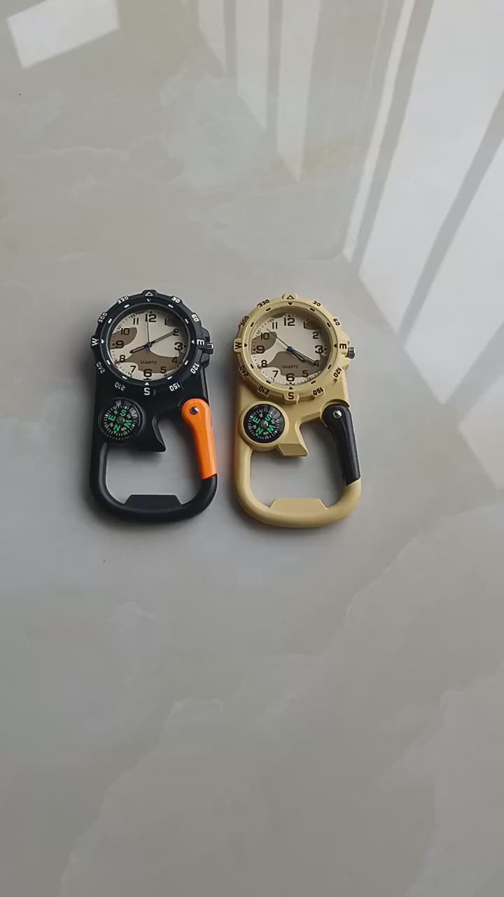 Black carabiner compass watch held in the hand: the watch face, the small compass and the carabiner clip, with a tan one beside it.