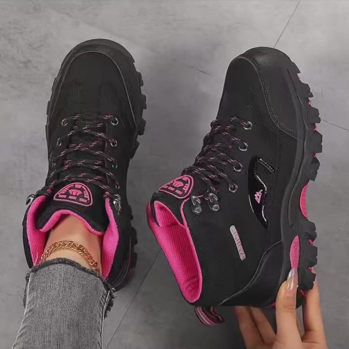 A pair of women's ankle hiking boots held up to the camera, black with rose-red trim, then the same boot in grey with rose red, showing the lug soles and padded collars.