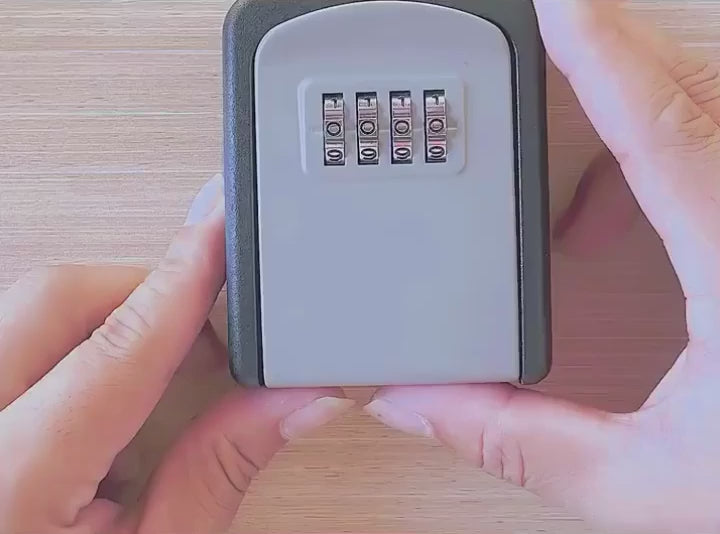 Hands resetting the code on the grey-face wall-mount key safe: the box opened, the lock panel lifted out, the dials turned to a new number, then the box closed.