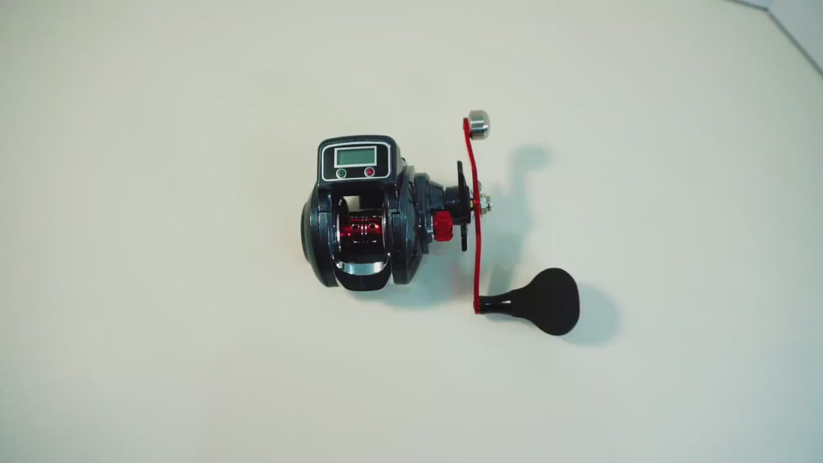 Dark grey and red low-profile reel with a digital line counter on the top plate, turned on a white background, then held in the hand while the counter's buttons are pressed.