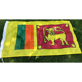 Sri Lankan flag in printed polyester laid flat on grass: gold lion with sword on maroon, green and orange bands, yellow edge