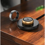 Gold R10 ring seated on the round black charging dock with its lead attached, silver ring beside it on a timber table