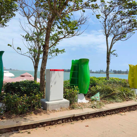 A green tulip turbine on a plinth beside a grey electrical cabinet in a coastal park, with more tulips along the shoreline path.