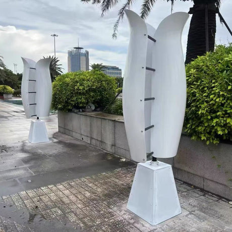 Two white tulip vertical turbines on tapered plinths on a wet paved plaza, with clipped hedges, palms and a city tower behind.
