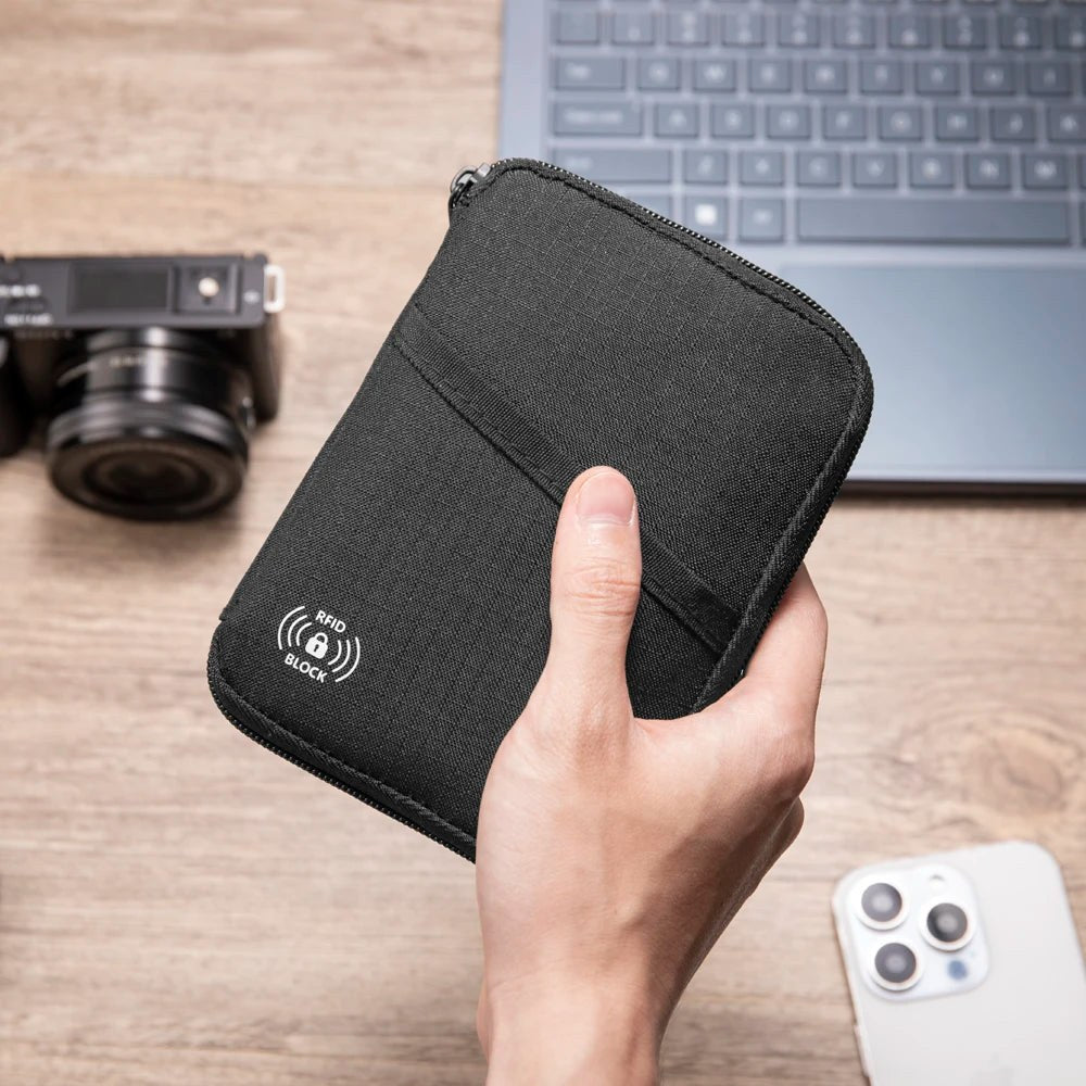 Black zip passport pouch held closed in one hand beside a laptop and camera